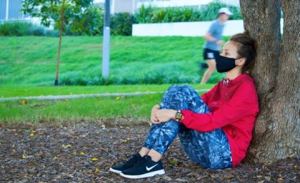 Woman sitting against tree with facemask on.